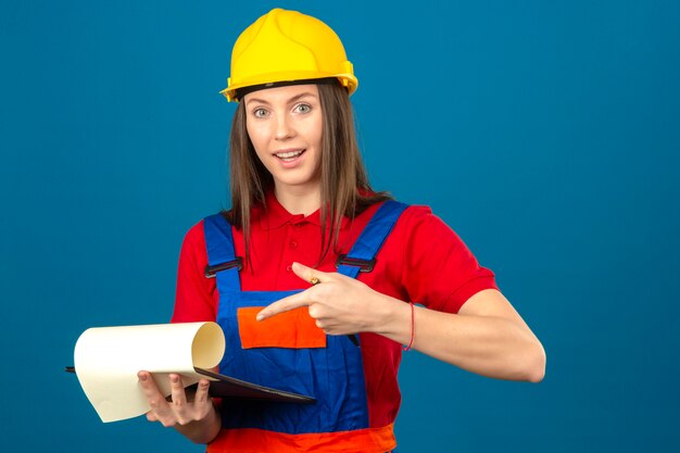 Jeune femme en uniforme de construction et casque de sécurité jaune souriant tenant le presse-papiers et pointant avec le doigt debout sur fond bleu