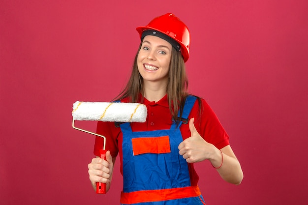 Jeune femme en uniforme de construction et casque de sécurité rouge souriant montrant signe ok et tenant le rouleau à peinture sur fond rose foncé
