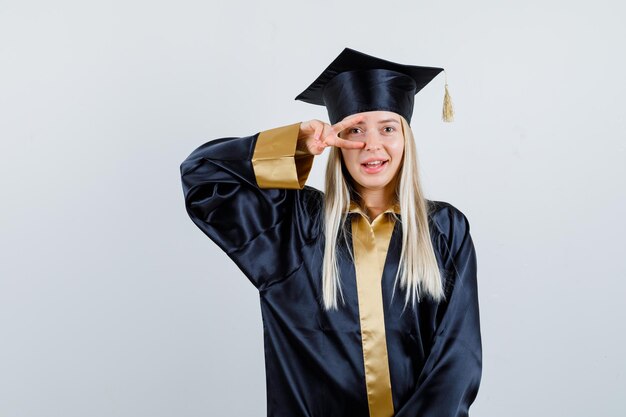 Jeune femme en uniforme diplômé montrant le signe V sur l'œil et l'air heureux