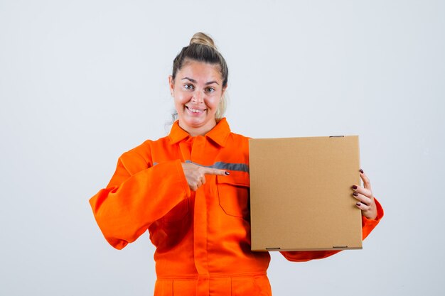 Jeune femme en uniforme de travailleur pointant sur la boîte et à la vue de face, heureux.