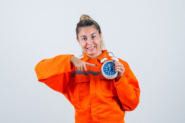 Jeune femme en uniforme de travailleur pointant sur l'horloge et à la vue de face, heureux.