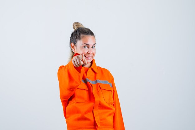 Jeune femme en uniforme de travailleur pointant vers l'avant et à la joyeuse vue de face.