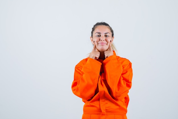 Jeune femme en uniforme de travailleur tenant les doigts sur ses joues et à la vue de face, adorable.