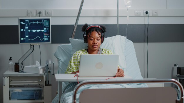 Jeune femme utilisant un casque et un ordinateur portable pour s'amuser à l'hôpital en attendant de guérir la maladie. Patient malade utilisant un casque pour écouter de la musique et se détendre au lit avec moniteur de fréquence cardiaque