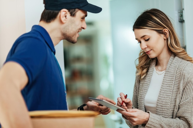 Jeune femme utilisant un pavé tactile pour signer la livraison d'un colis par un coursier tout en se tenant sur une porte