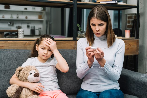 Jeune femme vérifiant la température sur le thermomètre assis près de la jeune fille assise avec ours en peluche