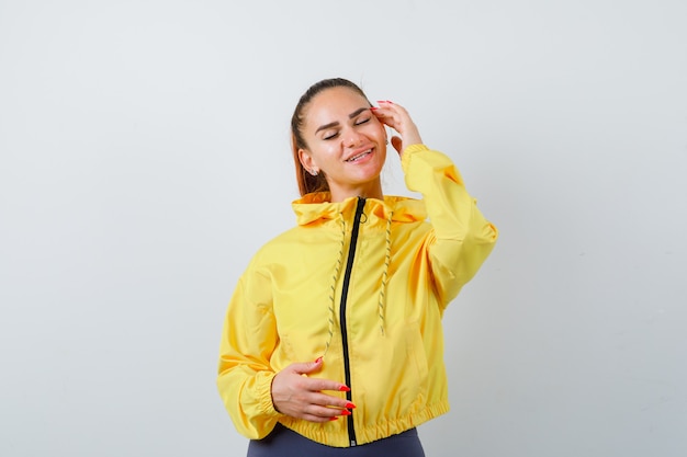 Jeune femme en veste jaune gardant la main sur la tempe, fermant les yeux et ayant l'air charmante, vue de face.