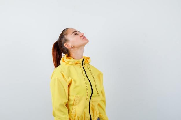 Jeune femme en veste jaune posant tout en regardant vers le haut et en ayant confiance en elle, vue de face.