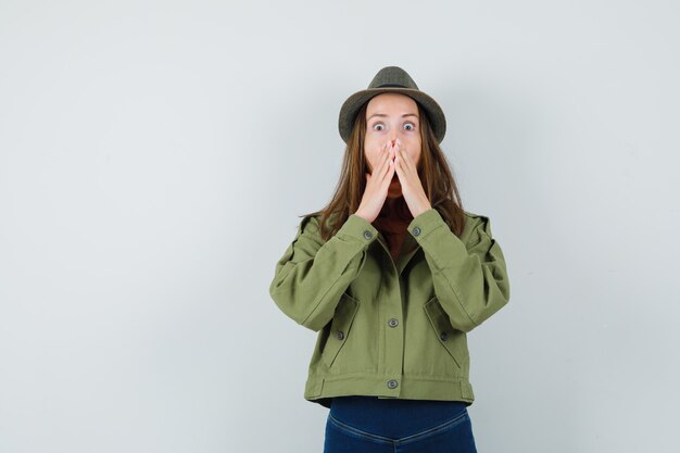 Jeune femme en veste, pantalon, chapeau tenant la main sur la bouche et à la surprise, vue de face.