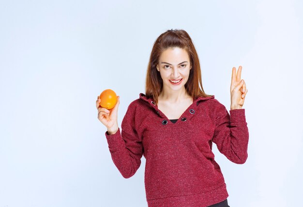 Jeune femme en veste rouge tenant une orange et montrant un signe positif de la main