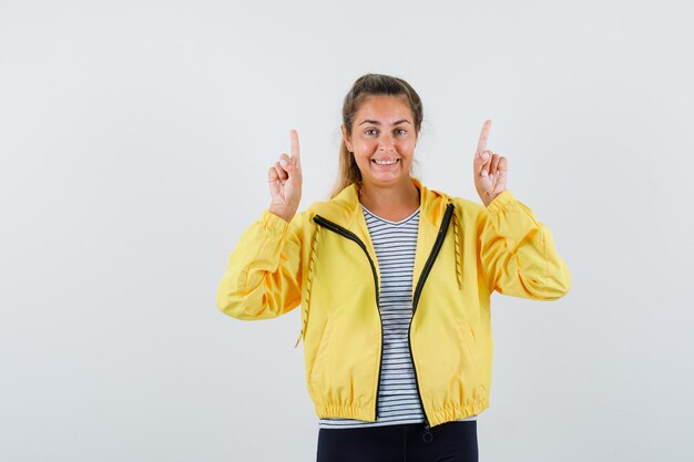 Jeune femme en veste, t-shirt pointant vers le haut et à la vue de face, joyeuse.