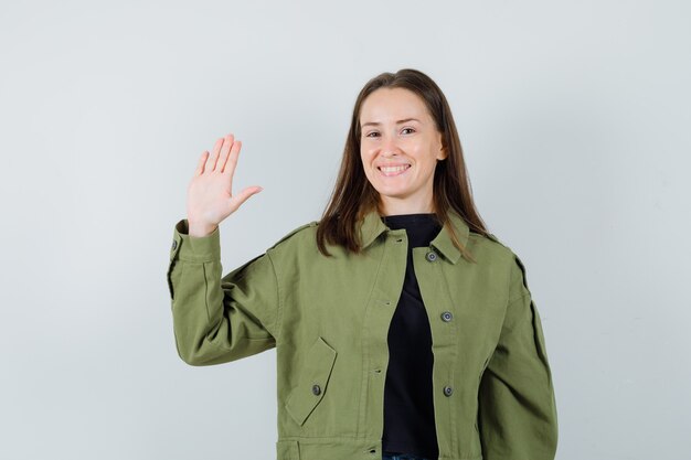 Jeune femme en veste verte en agitant la main pour saluer et à la vue de face, heureux.