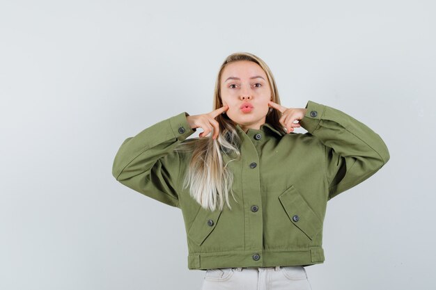 Jeune femme en veste verte, jeans pointant sur ses joues tout en faisant la moue des lèvres, vue de face.