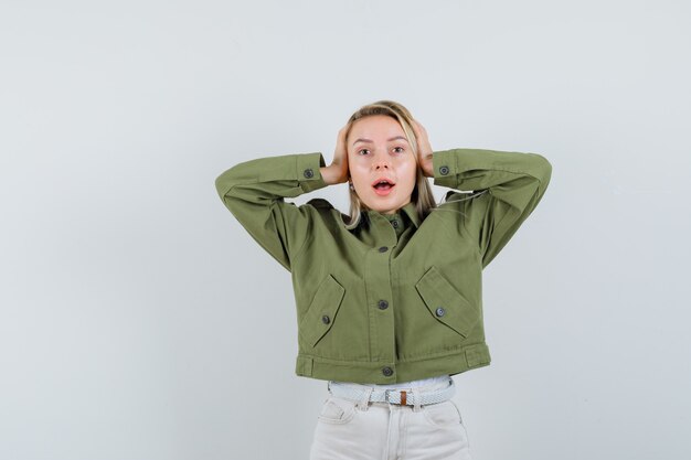 Jeune femme en veste verte, jeans tenant les mains sur l'oreille et à la vue de face, stressante.