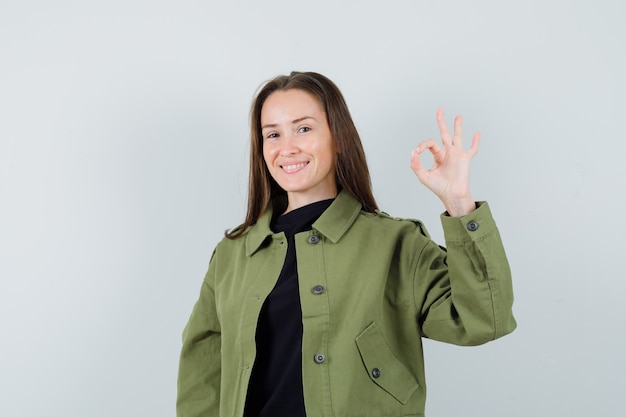 Jeune femme en veste verte montrant le geste ok et à l'optimiste, vue de face.