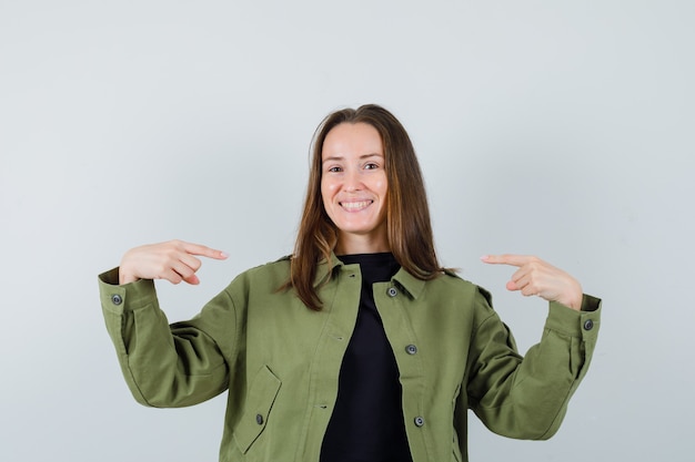 Jeune femme en veste verte pointant sur elle-même et regardant confiant, vue de face.