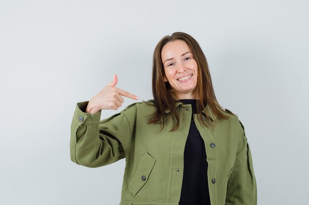 Jeune femme en veste verte pointant sur elle-même et regardant heureux, vue de face.