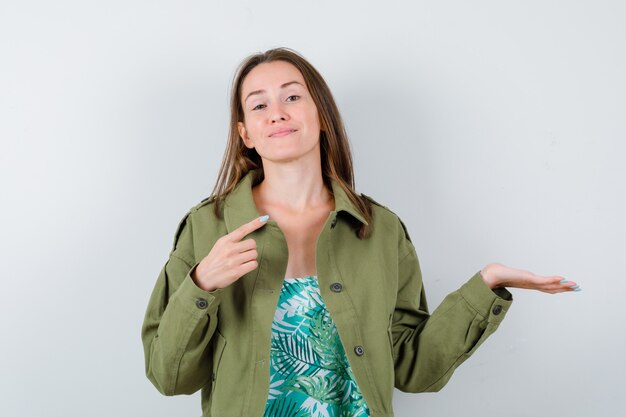 Jeune femme en veste verte pointant vers le coin supérieur droit, écartant la paume et l'air fière, vue de face.