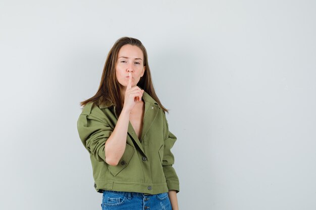 Jeune femme en veste verte, short montrant le geste de silence et regardant prudente, vue de face.