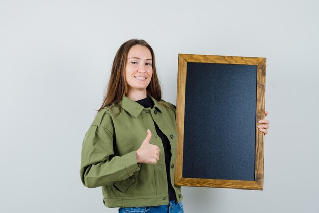 Jeune femme en veste verte tenant cadre noir tout en montrant le pouce vers le haut et à la vue de face, satisfait.