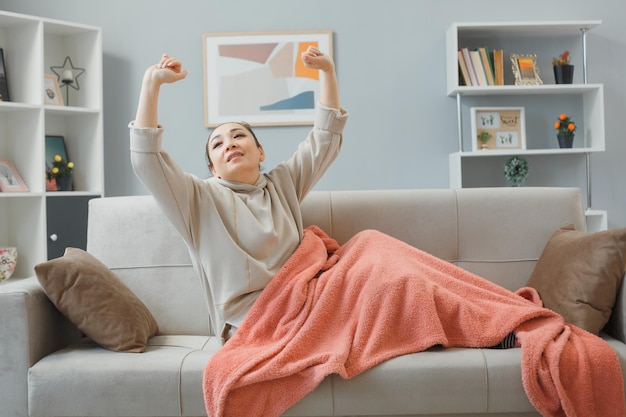 Jeune femme en vêtements de maison allant dormir sur un canapé recouvert d'une couverture à l'intérieur de la maison étant fatiguée après le travail en bâillant et en s'étirant se relaxant à la maison