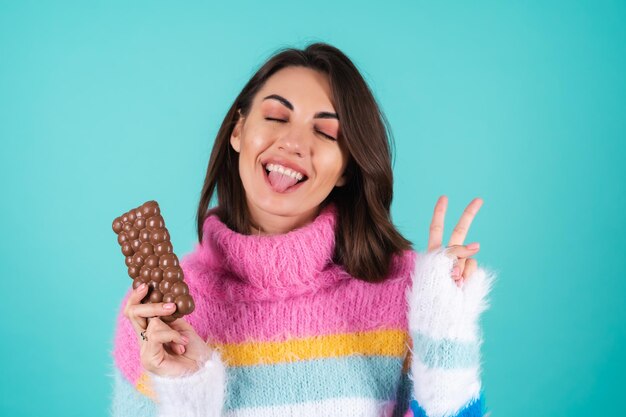 Une jeune femme vêtue d'un pull multicolore vif sur bleu apprécie chaque morceau d'une délicieuse barre de chocolat au lait aéré