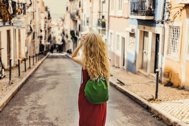 Jeune femme vêtue d'une robe rouge marchant dans une route entourée de bâtiments sous la lumière du soleil