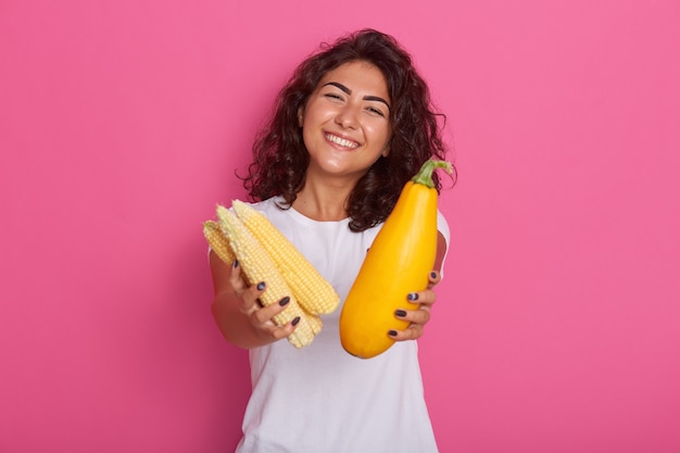 jeune femme vêtue d'un t-shirt décontracté blanc montrant des courgettes et des épis de maïs à la caméra, a une expression faciale heureuse, posant avec un sourire à pleines dents. Régime alimentaire cru et concept de saine alimentation.