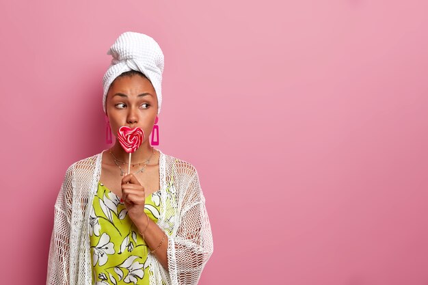 Jeune femme vêtue de vêtements décontractés, regarde sérieusement de côté, couvre la bouche avec une sucette en forme de coeur savoureuse sucrée, isolée sur un mur rose, espace copie vierge