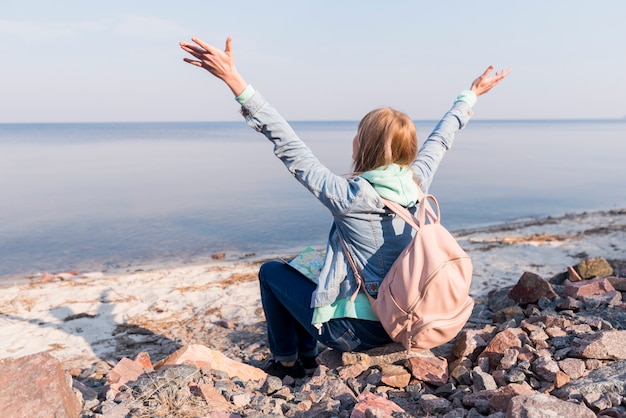 Jeune femme voyageur assis sur la plage en levant les bras surplombant la mer