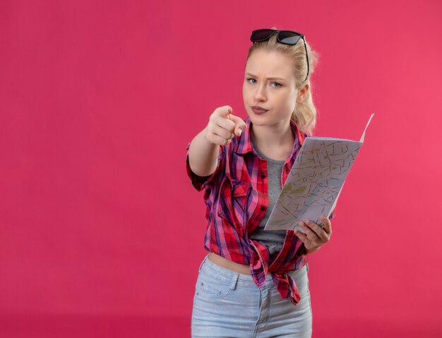Jeune femme voyageur portant chemise rouge et lunettes sur sa tête tenant la carte vous montre le geste sur un mur rose isolé