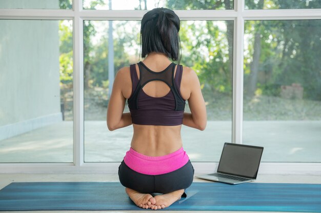 Jeune femme yogi pratiquant le yoga, faisant de l'exercice, assise, faisant de l'exercice, portant des vêtements de sport, regardant un didacticiel vidéo de remise en forme en ligne sur un ordinateur portable, faisant de l'exercice à la maison assis.