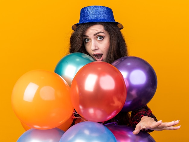 Jeune fêtarde impressionnée portant un chapeau de fête debout derrière des ballons regardant la caméra tendant la main isolée sur un mur orange