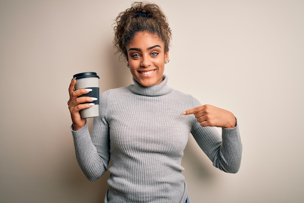 Jeune fille afro-américaine afro buvant une tasse de café à emporter sur fond blanc avec un visage surpris pointant du doigt vers lui-même