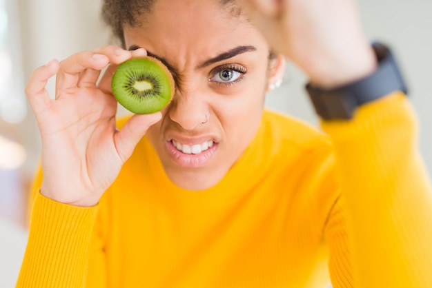 Photo gratuite jeune fille afro-américaine mangeant du kiwi vert agacée et frustrée criant de colère folle et criant avec la main levée concept de colère
