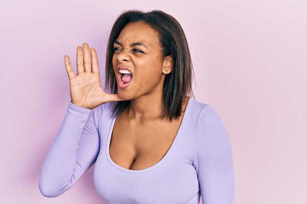 Jeune fille afro-américaine portant des vêtements décontractés criant et criant fort à côté avec la main sur la bouche. notion de communication.