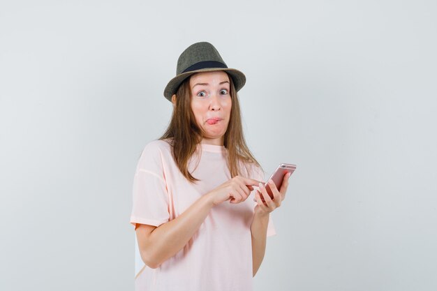 Jeune fille à l'aide de téléphone portable en t-shirt rose, chapeau et à la curieuse. vue de face.