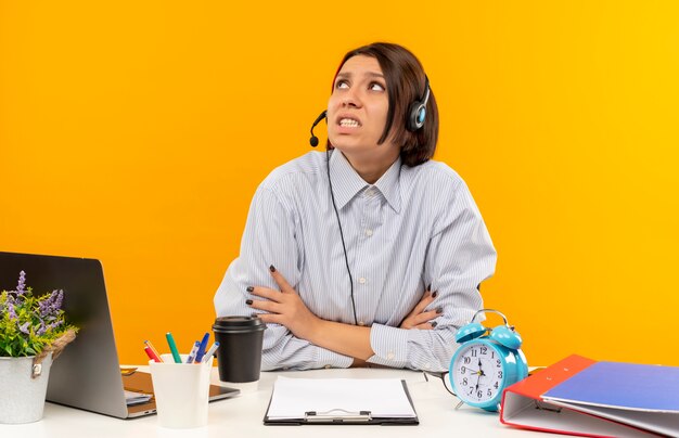 Jeune fille anxieuse de centre d'appels portant un casque assis avec une posture fermée au bureau en levant isolé sur orange