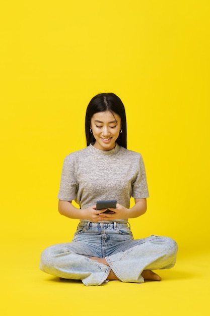 Jeune fille asiatique assise sur le sol tenant des textos de smartphone ou des achats en ligne isolés sur fond jaune Placement de produit pour la publicité d'application mobile Espace de copie