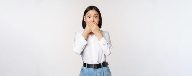 Une jeune fille asiatique choquée ferme la bouche, couvre les lèvres avec les mains et a l'air étonné debout sur fond blanc Geste tabou