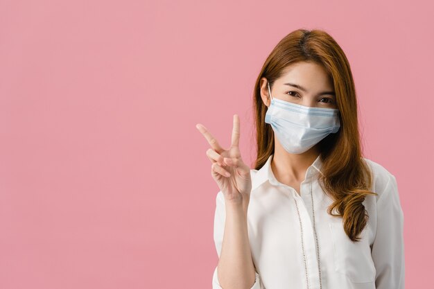 Jeune fille asiatique portant un masque médical montrant un signe de paix, encouragez avec vêtu d'un tissu décontracté et regardant la caméra isolée sur fond rose.