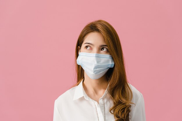 Jeune fille asiatique portant un masque médical vêtu d'un tissu décontracté et regardant un espace vide isolé sur fond rose.