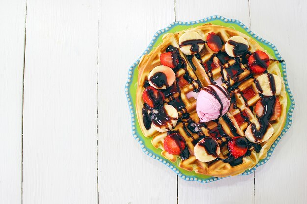 Jeune fille assise caffe en train de manger sa gaufre avec sa sauce au chocolat, ses tranches de banane et ses fraises sur une plaque en céramique verte et photographiée son petit-déjeuner