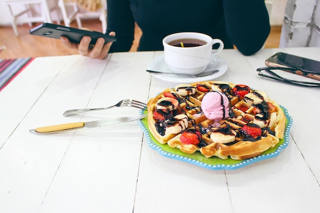 Jeune fille assise caffe en train de manger sa gaufre avec sa sauce au chocolat, ses tranches de banane et ses fraises sur une plaque en céramique verte et photographiée son petit-déjeuner