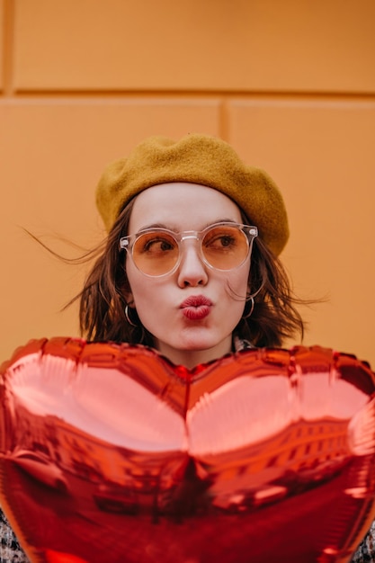 Jeune fille au béret jaune envoie un baiser Femme se cachant derrière un ballon en forme de coeur rouge