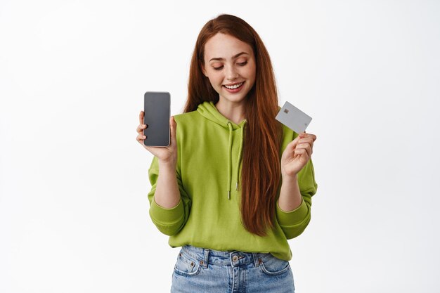 Jeune fille au gingembre avec un sourire blanc, regardant une carte de crédit, montrant un écran d'application de téléphone vide, introduisant une nouvelle interface, une application sur un téléphone portable, un fond blanc.