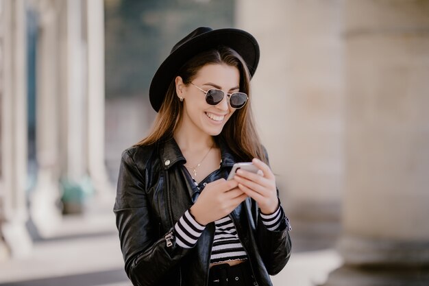 Jeune fille aux cheveux bruns dans une veste en cuir, chapeau noir sur la promenade de la ville et joue sur téléphone mobile