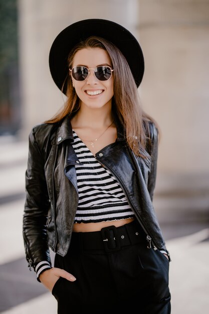 Jeune fille aux cheveux bruns dans une veste en cuir et des lunettes, chapeau noir sur la promenade de la ville