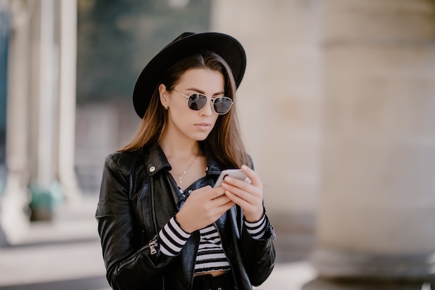 Jeune fille aux cheveux bruns dans une veste en cuir et des lunettes, chapeau noir sur la promenade de la ville