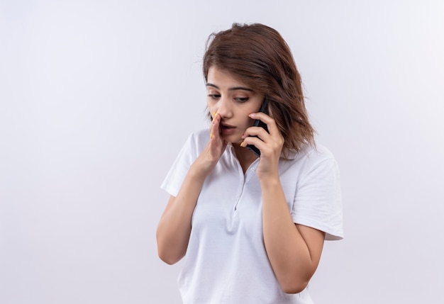 Jeune fille aux cheveux courts portant un polo blanc chuchotant en disant un secret tout en parlant au téléphone mobile